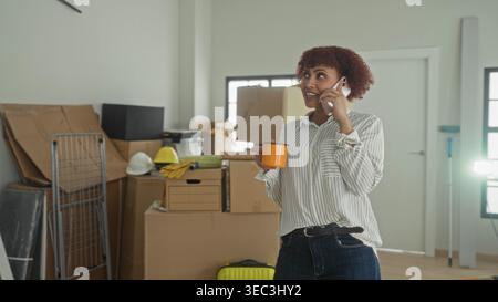Woman talks on smartphone while holding an orange coffee mug, smiling amid packed boxes, ladder and moving gear inside a bright building during a new Stock Photo