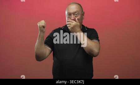 Hispanic senior man covers mouth with hand, clenches fist and looks startled in red studio; surprise silence. Stock Photo