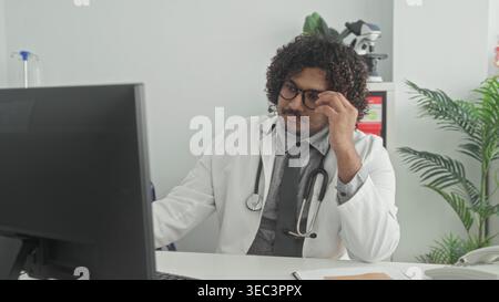 Man doctor in white coat holds eyeglasses and rubs his eyes at a clinic desk in a building; quiet fatigue. Stock Photo