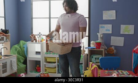 Man picks up basket and bends to pick toys, hand holding wicker basket visible among colorful toys, shelves and play tent in kindergarten building; pa Stock Photo