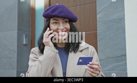 Woman on phone holding creditcard at building entrance near intercom and door, wearing purple beret and beige coat while checking card; payment verifi Stock Photo