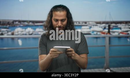 Man clenches fist holding smartphone at port railing near moored boats, eyes closed and gripping device; gaming victory joy. Stock Photo