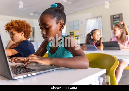 Diverse classmates with African American girl typing on laptop at classroom table with tablets Stock Photo