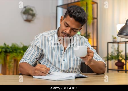 Indian man at home drinks coffee and writes goals in notebook ticks boxes and plans day with drive. Arabian guy at desk reviews list adds tasks sets priorities smiles feels focus and motivation aim Stock Photo