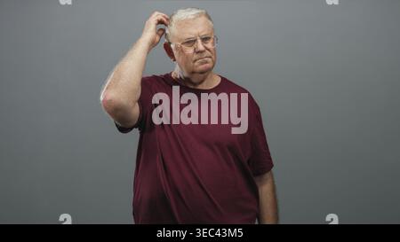 Senior man in burgundy t shirt scratching head with right hand while wearing glasses in studio grey wall setting; confusion reflection pondering. Stock Photo