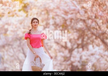 Portrait of a young woman standing in front of flowering sakura trees in springtime holding a straw basket, Thailand Stock Photo