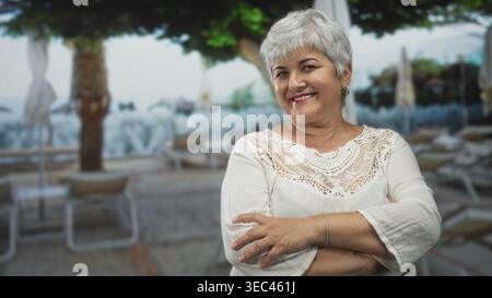 Woman with arms crossed smiling, white lace top and bracelet on a seaside street with loungers and umbrellas; contentment relaxation. Stock Photo