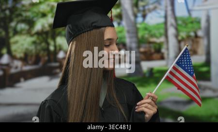 Woman holding flag in park, smiling in black graduation cap and gown with red tassel, standing on green lawn by benches; pride. Stock Photo