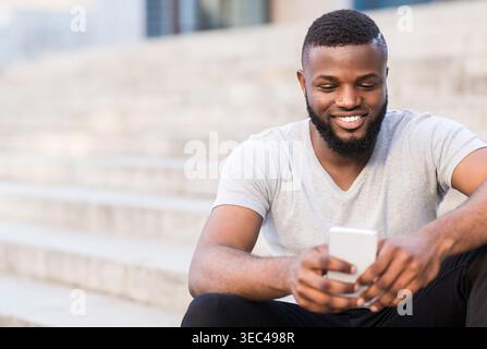 Young Man Sitting on Steps Using Smartphone Outdoors While Smiling in a Casual Setting Stock Photo