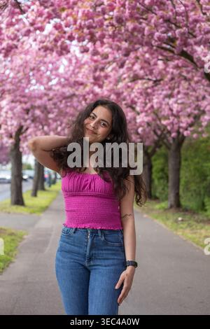 Smiling young woman posing on street with cherry blossom trees in spring Stock Photo
