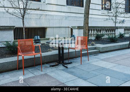 Empty outdoor seating area with two orange chairs and a round table near a building in the city during daytime hours Stock Photo