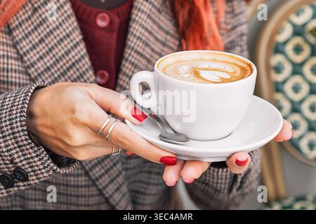 Woman in plaid blazer holding a warm cup of cappuccino with latte art, enjoying a relaxing moment in a cafe Stock Photo