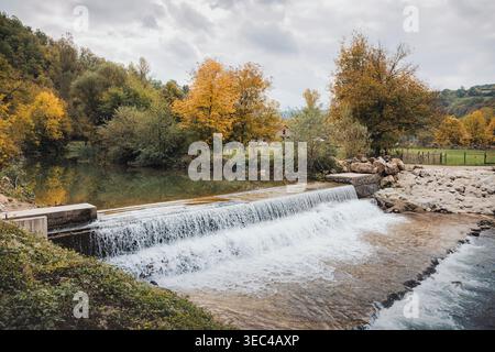 River water creating a small waterfall over a man made weir, surrounded by colorful autumn trees and a rural landscape Stock Photo