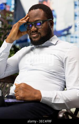 African american business person fatigued and distressed due to bad news read on his laptop, struggling to work remotely outdoor on a bench. Surrounded by urban metropolitan life. Stock Photo