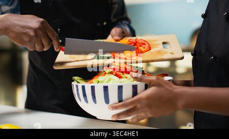 Black couple in aprons adding vegetables in a bowl and mixing ingredients, making a fresh vegetarian salad for their lunch meal. Healthy lifestyle and positive relationship. Stock Photo
