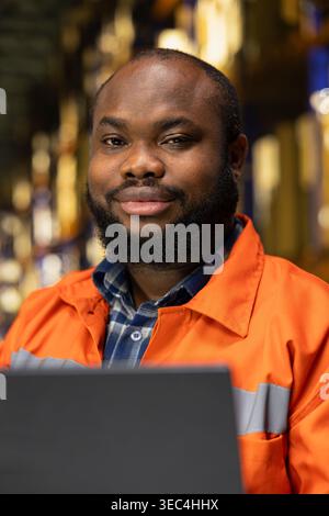 Portrait of black man using laptop for supply chain management, coordinating import export shipment and parcel tracking for e-commerce operations. Manual labor for large business facility. Stock Photo