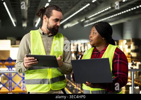 Two diverse workers, collaborating on logistics, tasks for e-commerce, logistics and order fulfillment with laptop and tablet on platform. Shipping operations productivity, supply chain. Stock Photo