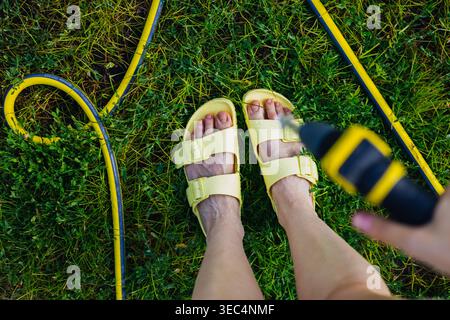 Woman in yellow sandals spraying water on her feet with a garden hose to cool down. Concept of summer heat relief, sensory wellness, and backyard fun. Stock Photo