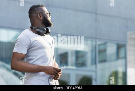 Man Wearing Headphones Stands Outside Urban Building Enjoying Music on a Sunny Day Stock Photo