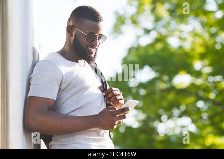 Young Man Using Smartphone While Leaning Against Wall in Bright Outdoor Setting Stock Photo