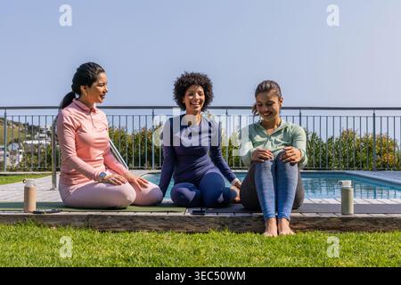 Diverse female friends sitting on yoga mats at poolside deck, in athletic wear with water bottles Stock Photo