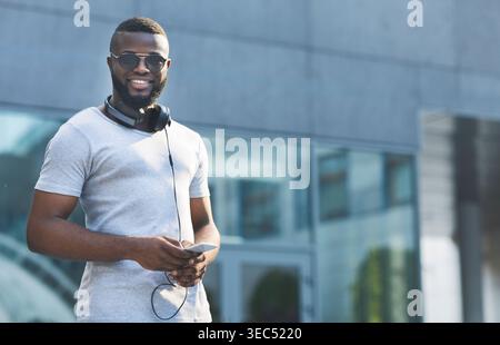 Young Man Stands Confidently Outside a Modern Building Using Smartphone With Headphones Stock Photo
