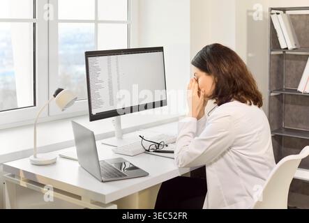 Tired female doctor with headache rubbing eyes at computer in bright clinic office Stock Photo