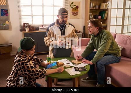 Young adult roommates budgeting shared expenses, counting cash, checking smartphone banking, writing notes at table. Ideal for student finance, rent splitting, cost of living content Stock Photo