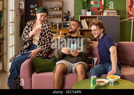 Young adult students sharing smartphone content while sitting together in apartment, laughing and socializing. Ideal for student lifestyle, friendship, campus housing, digital communication Stock Photo