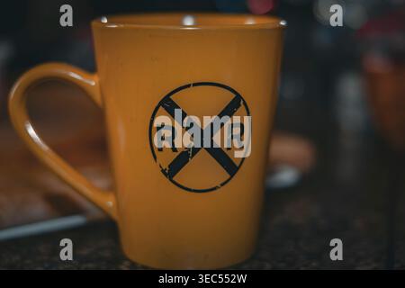 Close up shot of a bright yellow coffee mug featuring a weathered black railroad crossing symbol RXR against dark moody background on granite counter Stock Photo