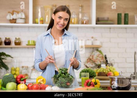 Young Woman Prepares Fresh Salad in Bright Kitchen With Various Vegetables on the Counter Stock Photo