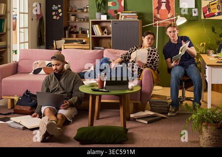 Wide shot of diverse students studying in shared apartment, using laptop and books while discussing coursework. Ideal for education marketing, student housing, campus life content Stock Photo