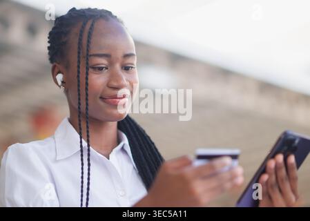 Young Black business woman using smartphone and credit card at outdoor cafe table with tablet nearby. Online payment, mobile banking, digital commerce Stock Photo