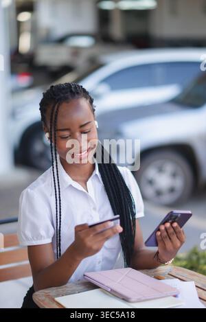 Young Black professional woman using smartphone and credit card while sitting at cafe table with tablet. Mobile payment, online banking, fintech comme Stock Photo