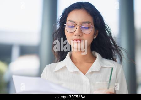 Focused young professional reading printed documents while holding iced coffee outside a modern office building, showing concentration, corporate rout Stock Photo