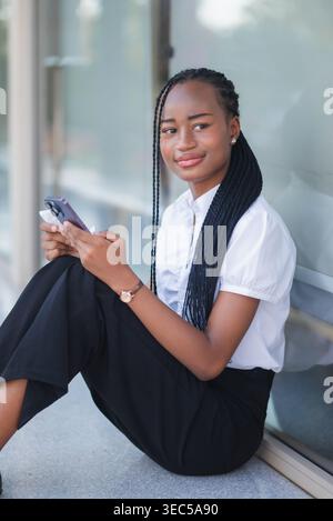 Young Black business woman seated outdoors with smartphone and paper receipt, looking thoughtful. Purchase verification, payment checking, budget revi Stock Photo