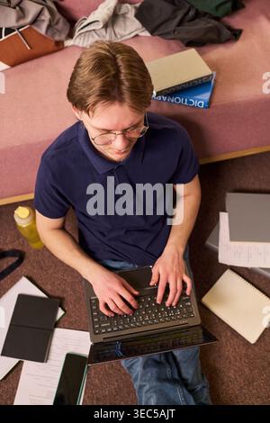Vertical shot of Caucasian male student typing on laptop while sitting on floor beside study materials, preparing coursework. Useful for education marketing, campus life, online learning Stock Photo