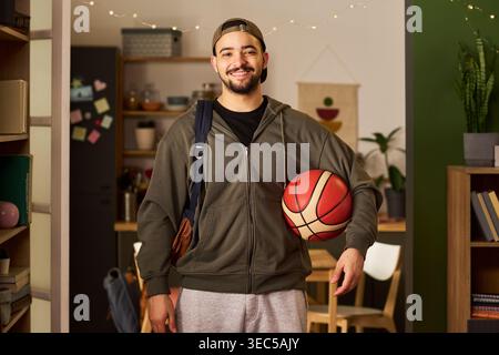 Portrait of young adult biracial man carrying basketball and bag, smiling at camera in home interior, representing student lifestyle and active routine. Useful for education or youth marketing Stock Photo
