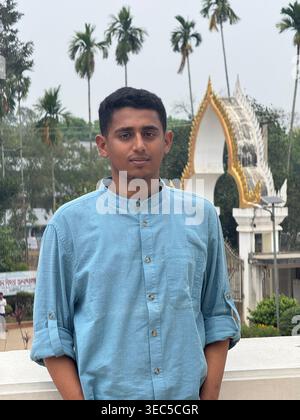 A stylish young man stands confidently at a scenic temple entrance, surrounded by tall palm trees and a peaceful natural backdrop. Wearing sunglasses Stock Photo