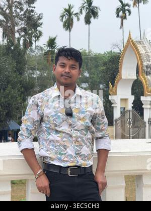 A stylish young man stands confidently at a scenic temple entrance, surrounded by tall palm trees and a peaceful natural backdrop. Wearing sunglasses Stock Photo