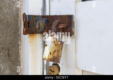 rusty metal padlock and hasp on weathered white wooden door, corroded brown surfaces side view in soft focus closeup, security and urban decay concept Stock Photo