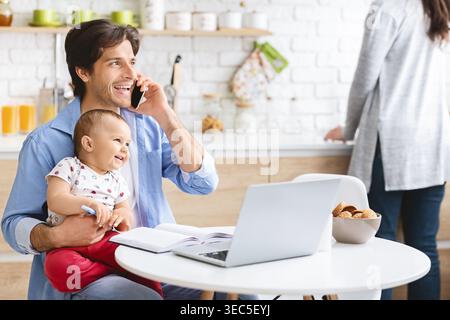 Happy Father Talking on the Phone While Holding Baby in a Modern Kitchen Stock Photo