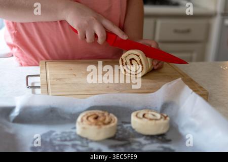 Close-up of hands cutting cinnamon roll dough on wooden board with raw rolls on baking tray, home baking process in kitchen Stock Photo