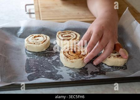 Close-up of hand placing raw cinnamon rolls on baking tray with parchment paper, homemade baking process in kitchen Stock Photo