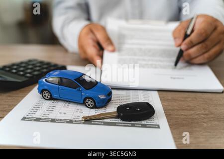 Hands reviewing car loan finance document contract with payment schedule table, blue toy car model and key on paperwork, calculator on desk, represent Stock Photo