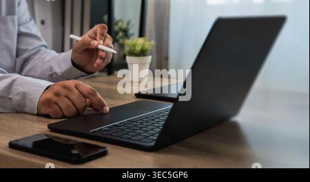 Hands using laptop keyboard and stylus on desk with smartphone and plant, interacting with touchpad and typing workflow for digital communication and Stock Photo