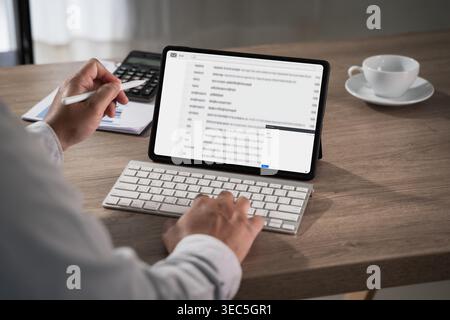Hands using tablet displaying email inbox with keyboard on desk alongside calculator document and coffee cup, managing message communication workflow Stock Photo