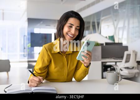 Businesswoman sits at desk, checking smartphone, taking notes in notebook Stock Photo