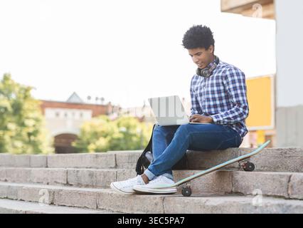 Young Person Working on Laptop While Sitting on Steps With Skateboard Nearby at Outdoor Location Stock Photo