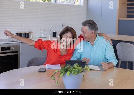 Senior woman in red blouse and man in blue shirt sitting at kitchen table using tablet Stock Photo